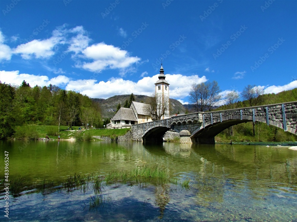 Beautiful view of the St. John the Baptist church next to Bohinj lake next to a arched stone bridge and a reflection in the lake and white clouds in blue sky