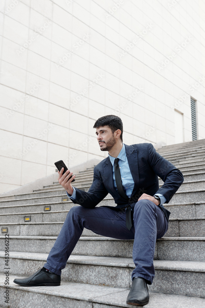 Confident young businessman sitting on steps outdoors and dialing number of colleague