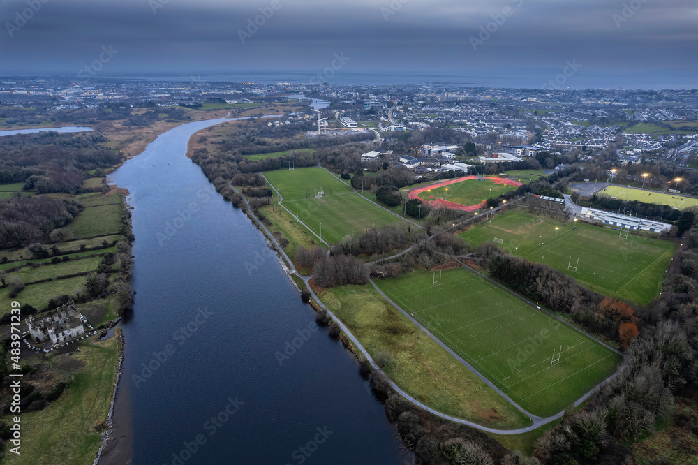 River Corrib flows into Galway city, aerial view. Sport ground with tall goal posts for Irish national sport on the right. Training field for hurling, rugby, camogie, Gaelic football. Dusk time.