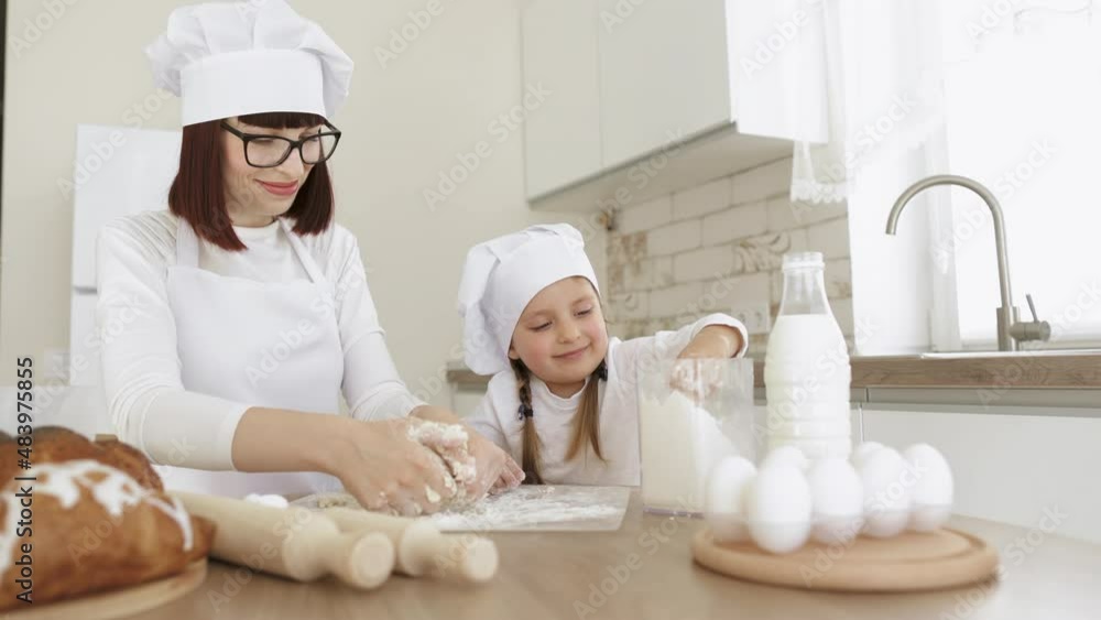 Charming adorable attractive beautiful caucasian sprinkle flour on the dough while smiling to young mom in hats of chef and white apron for cooking. Family in a kitchen. Charming adorable attractive beautiful caucasian sprinkle flour on the dough while smiling to young mom in hats of chef and white apron for cooking. Family in a kitchen.