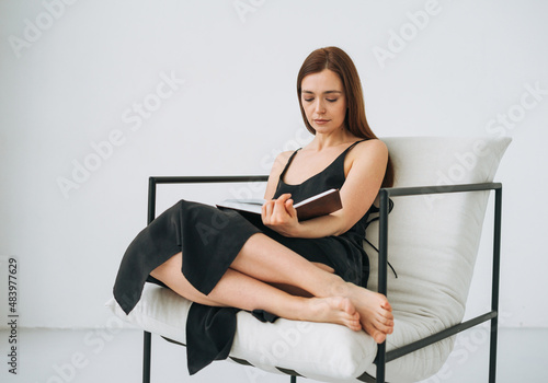 Beautiful young woman with dark long hair in black dress reading book on chair in the white interior