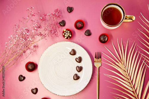 Chocolate hearts and decorated sweets. Romantic dinner, Valentines Day concept. White mock up vintage plate with fork and cup of tea. Pink background, gypsophila and golden decorations. Top view