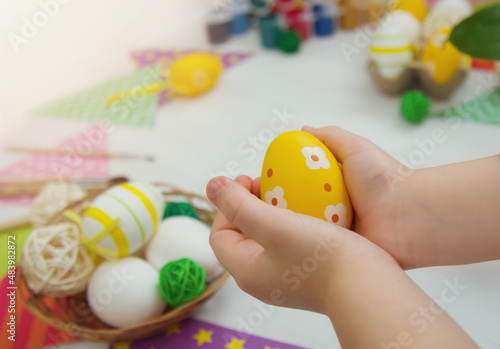 child is holding a yellow Easter egg with a pattern. Children's creativity, master class. Hands of a baby with an egg close-up against the background of a basket with eggs, paints, brushes.