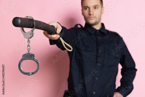 One handsome policeman officer wearing black uniform with club and handcuffs isolated on pink background. photo