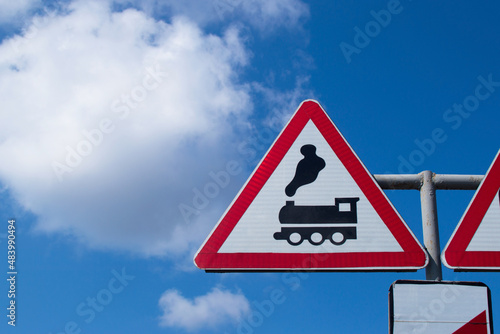 Railroad Level Crossing Sign without barrier. A road sign depicting an old black steam locomotive in a red triangle, A bright warning sign on a pole against a blue sky.