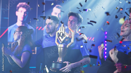 Happy professional esport team of gamers winners with golden cup standing under falling confetti and celebrating victory in professional esports gaming tournament together photo