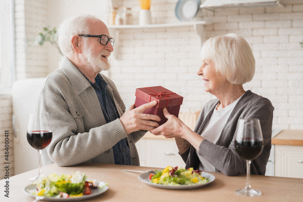 Caucasian senior elderly old couple spouses grandparents giving present gift on romantic date at dinner at home, drinking wine together on anniversary Valentine`s Day Caucasian senior elderly old couple spouses grandparents giving present gift on romantic date at dinner at home, drinking wine together on anniversary Valentine`s Day