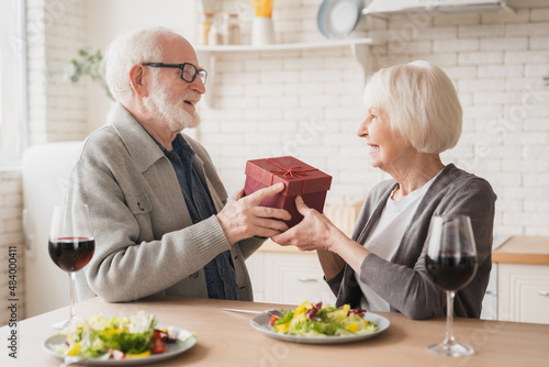 Caucasian senior elderly old couple spouses grandparents giving present gift on romantic date at dinner at home, drinking wine together on anniversary Valentine`s Day