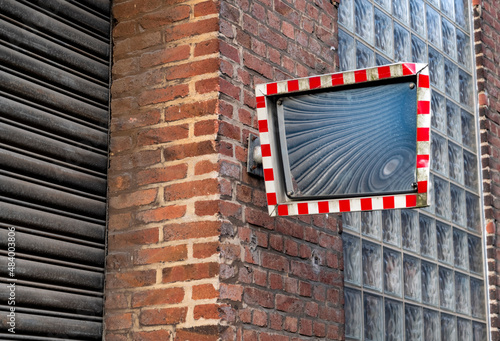 Dented old traffic mirror made of polished metal with red and white chequered frame on the red brick facade of a ruinded factory building in Germany. Rolling gate and glass building blocks. photo