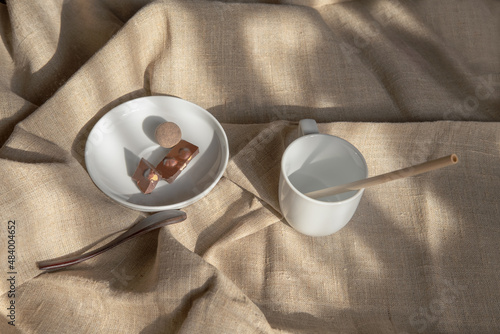 White cup with a bamboo tube and a saucer with chocolate and a round candy on a light textile background in soft rays of light