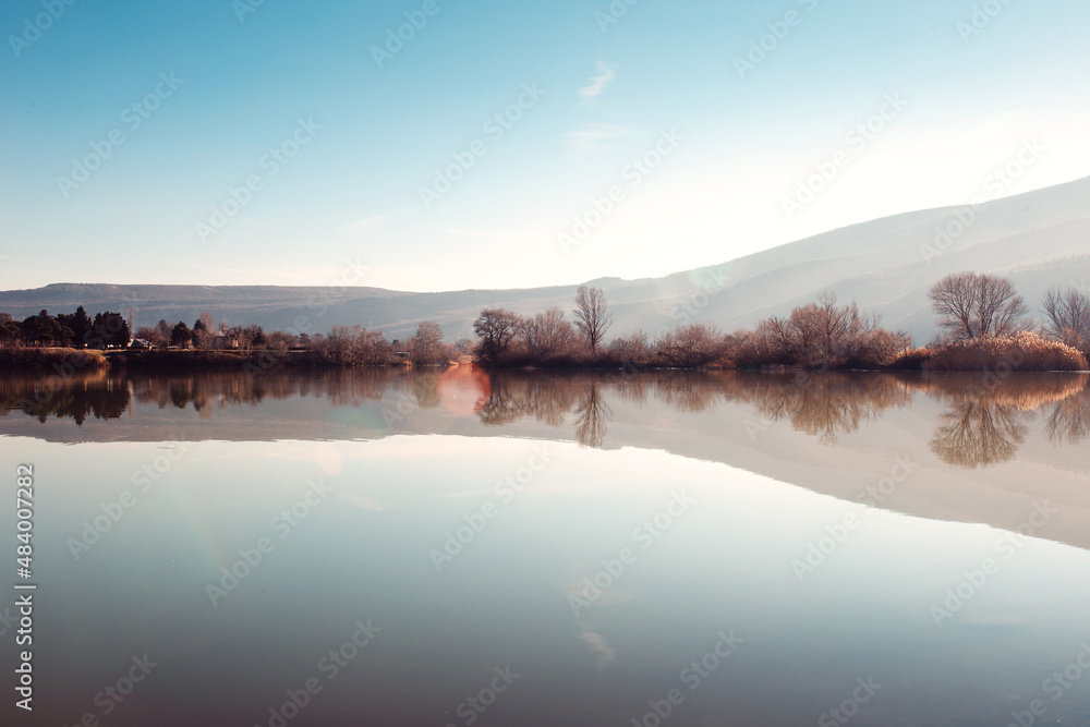 Dawn over the lake with a quiet smooth surface of the water and the reflection of mountains and trees in it - a peaceful landscape in the rays of the low sun