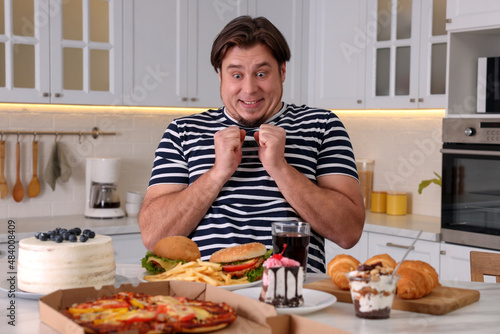 Hungry overweight man at table with sweets and fast food in kitchen