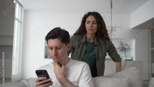 Young woman looks out over husband shoulder at phone screen and starts quarreling accusing man in cheating in living room photo
