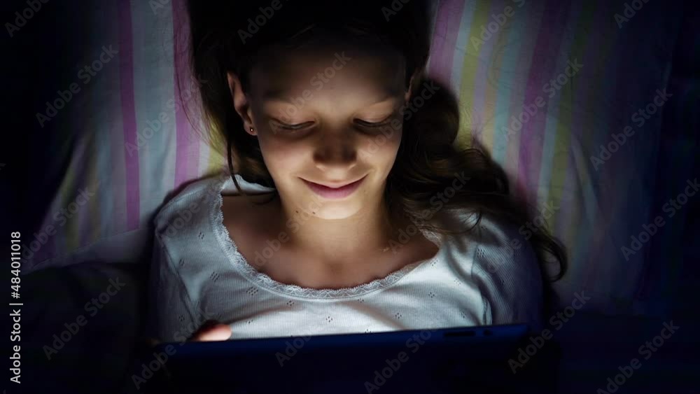 Girl lying in bed at night and watching funny video on tablet, playing online game, browsing internet, light from screen on her face, bedtime with gadget, addicted kid, top view. Concept of technology Girl lying in bed at night and watching funny video on tablet, playing online game, browsing internet, light from screen on her face, bedtime with gadget, addicted kid, top view. Concept of technology