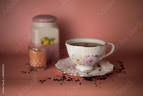 a white and pink cup with flowers with black tea and a ceramic tea jar and scattered tea and a burning pink candle in a glass with pearls on a pink background