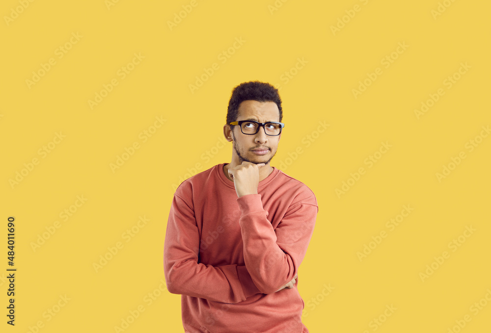 Fototapeta premium Hmm. Young student in glasses doubting something. Puzzled adult man in casual sweatshirt isolated on solid yellow background looking up and thinking hard about difficult question or future career plan