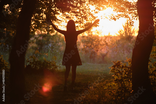 Dark silhouette of young woman walking alone through dark forest joyfully raising her hands in summer evening. Enjoying nature and outdoor activities concept