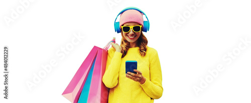 Colorful portrait of stylish smiling young woman listening to music in headphones with shopping bags posing wearing a yellow knitted sweater, pink hat isolated on white background