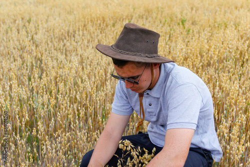 Harvester. Portrait of farmer seating in gold wheat field with blue sky in background. Young man wearing sunglasses and cowboy hat in field examining wheat crop. Oats grain industry. Closeup