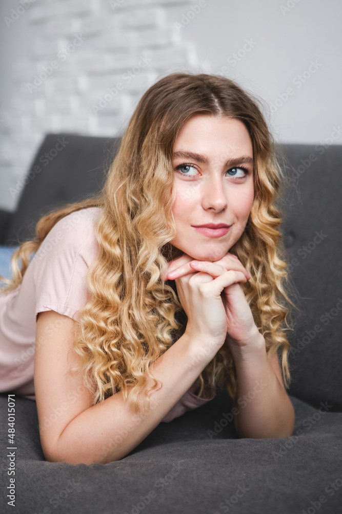 Beautiful young woman sitting on gray sofa in the room. Resting girl in pink shirt. Relaxing woman Beautiful young woman sitting on gray sofa in the room. Resting girl in pink shirt. Relaxing woman