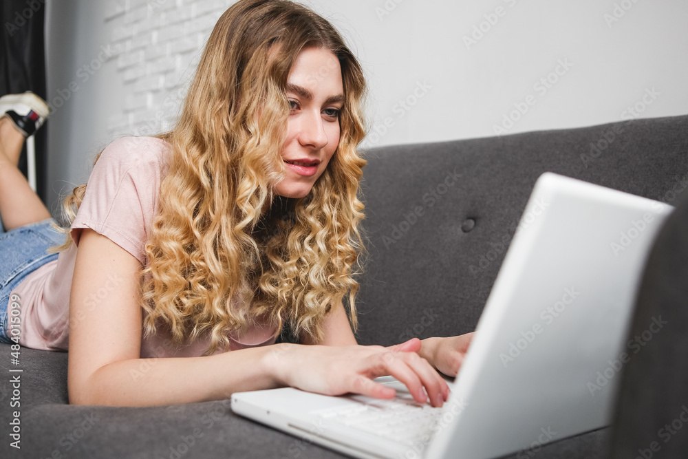 Beautiful young woman in pink shirt working on the laptop. Girl lying on the sofa with notebook. Distance learning student Beautiful young woman in pink shirt working on the laptop. Girl lying on the sofa with notebook. Distance learning student