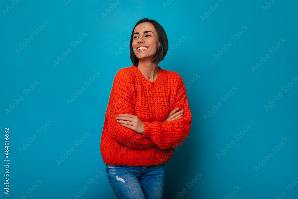 Fototapeta premium Gorgeous smiling young brunette woman in bright color sweater is posing with crossed arms on blue background and looking away