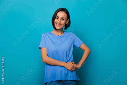 Close up portrait of happy beautiful young brunette woman with stylish hairstyle in blue t-shirt while she is posing and isolated on blue background in studio
