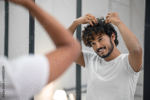 Pleased focused guy styling his curly hair with foaming mousse