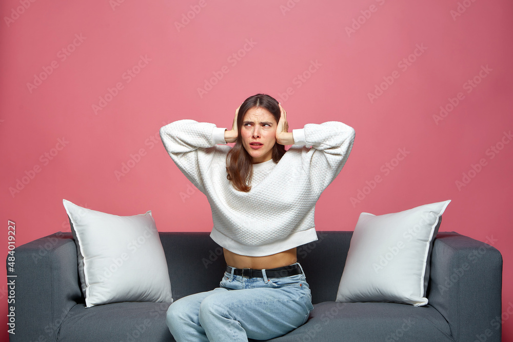 Annoyed stressed young girl cover ears suffer from loud noise sound, sitting on sofa, not listen noisy music, neighbors Annoyed stressed young girl cover ears suffer from loud noise sound, sitting on sofa, not listen noisy music, neighbors