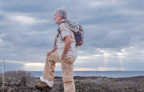 Adult caucasian gray haired senior man hiking in countryside close to the sea wearing backpack looking at the horizon enjoying nature. Freedom and vacation concept. Cloudy sky. Copy space
