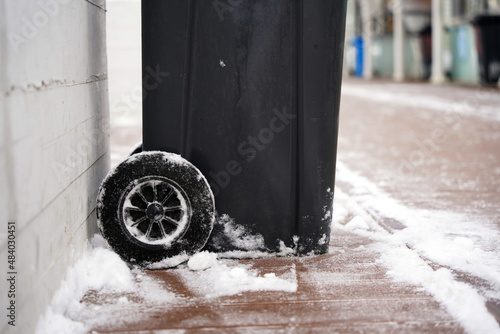 Black plastic garbage can with wheels on a snow-covered sidewalk photo