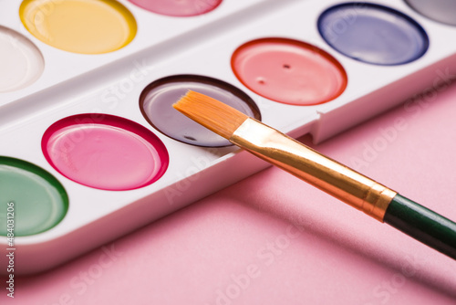 Multicolored watercolor paints with a brush on a pink background. Objects for art