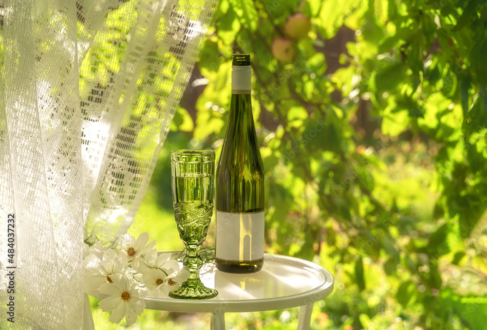 Beautiful vintage glass and bottle of white wine with white flowers on glass table in summer garden in sunlight. Copy space. Summer drinks concept.
