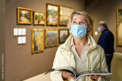 elderly woman in mask protecting against covid examines paintings on display in hall of art museum photo