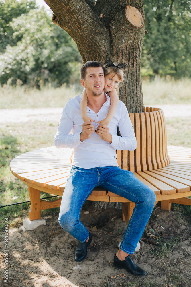 Fototapeta premium Dad and daughter sitting on a bench under a tree. Little girl in a black dress hugs dad. Spend time with family in the park. Father's Day