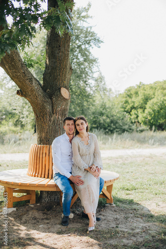 man and woman sitting on a bench under a tree. Beautiful brunette in a long beige dress. Married couple on a walk. happy couple