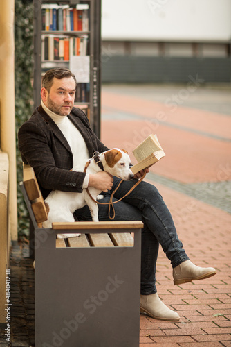 A man reading a book on a bench in the city square with a dog in the city square with a dog. Jack Russell terrier and owner. Pets photo