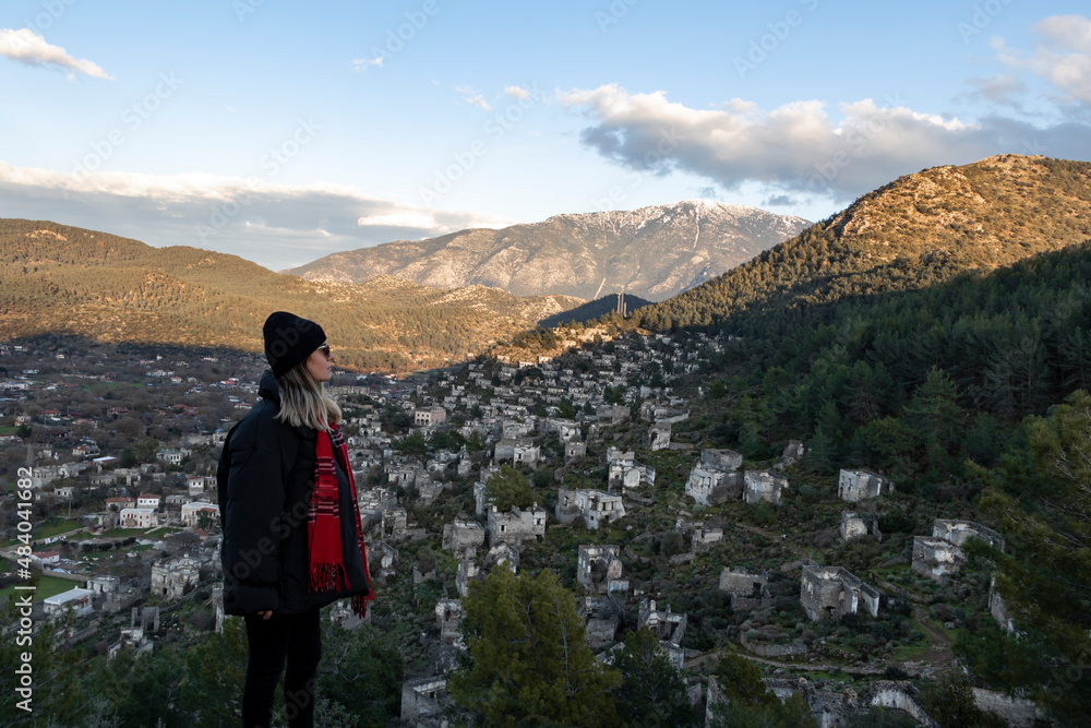 Girl is on the top of the mountain. She has black hat. Watching amazing abandoned city view. Cloudy sky, mountains background. Adventure and travel concept photo.