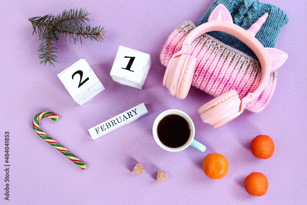 Obraz premium Calendar for February 21: the name of the month February in English, the numbers 21, a warm hat, headphones, a cup of coffee, sugar cubes, fruits and candies, pastel background, top view