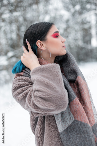Stylish beautiful young woman model with bright colored makeup and pink eye shadows in a fashionable fur coat and scarf walks in the snow in the park in the woods.