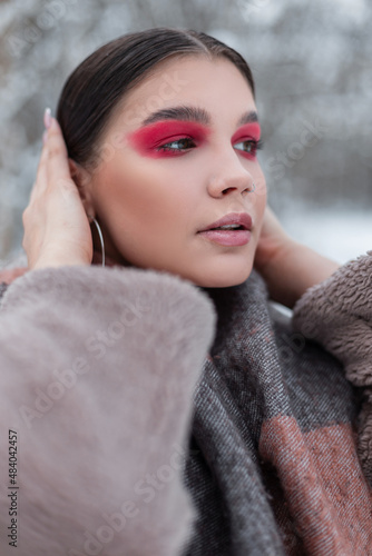 Fashion portrait of a beautiful young stylish woman with bright pink makeup in winter fashionable clothes with a scarf walks on a winter day in a snowy park