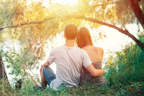 Couple boy and girl teenagers holding hands. The guy loves the girl. Young lovers sit on the shore of the lake near the tree.