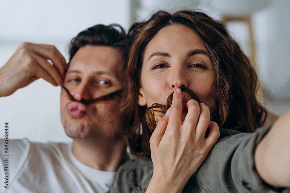 Happy newlyweds play and pose making mustaches with curly brunette hair and kiss smiling broadly for selfies via smartphone closeup Happy newlyweds play and pose making mustaches with curly brunette hair and kiss smiling broadly for selfies via smartphone closeup