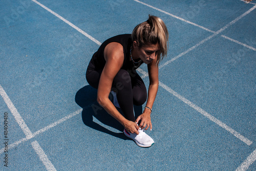 Woman on the running track tying the laces of her sneakers. Athletic training. Blue running track. Woman in black sportswear. © TxemaPhoto