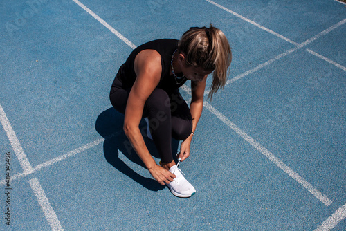 Woman on the running track tying the laces of her sneakers. Athletic training. Blue running track. Woman in black sportswear.