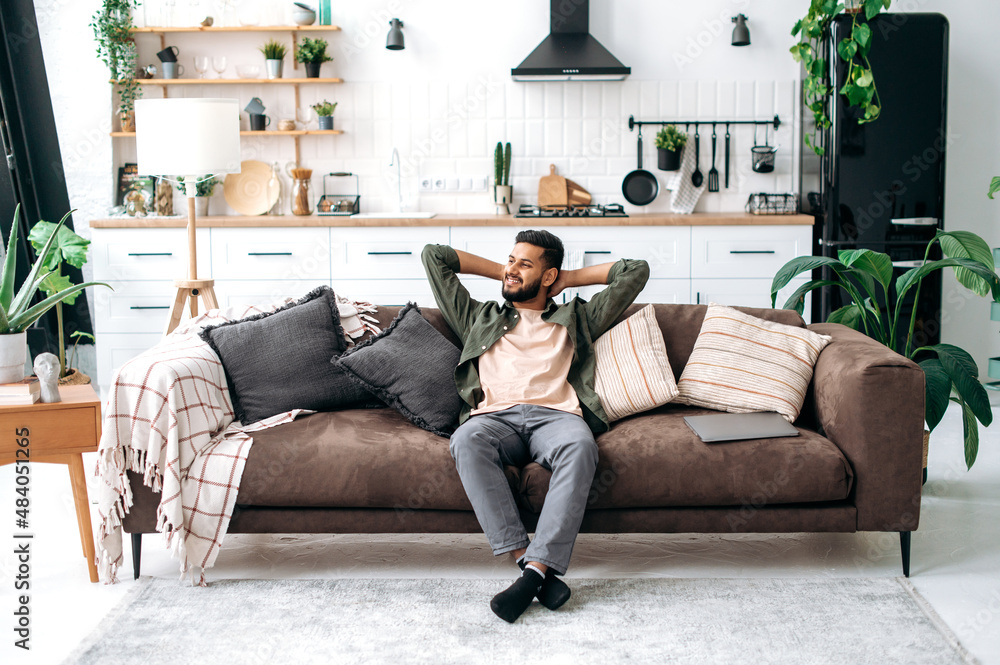 © Kateryna - Satisfied relaxed young indian man chilling on sofa in living room, smiling. Handsome arabian guy have rest at home, after laptop distant work, dreaming about vacations, throwing his hands behind head