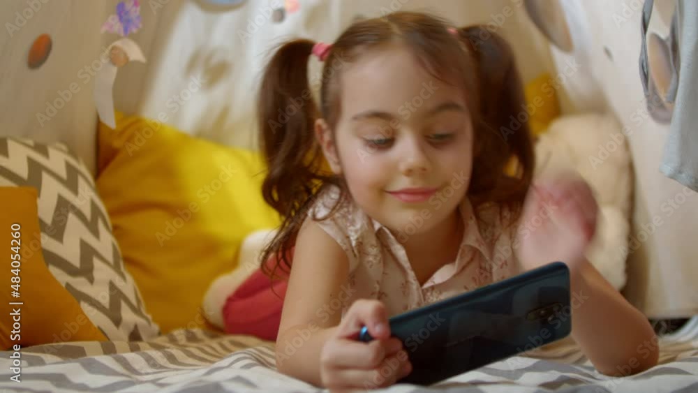 Cute smiling preschool girl watches cartoons on her smartphone, plays online games, has fun on vacation at home in a tent. Cute smiling preschool girl watches cartoons on her smartphone, plays online games, has fun on vacation at home in a tent.