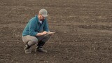 Farmer enters data for report on soil condition into tablet. Man agronomist squats on black ground plot of plowed and cultivated field in countryside on spring day. Agricultural business concept