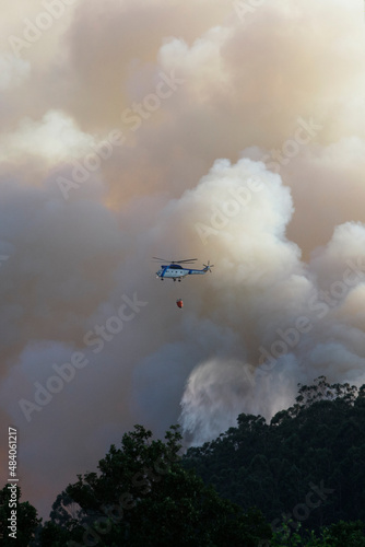 A helicopter flies by discharging water amid clouds of smoke from a forest fire