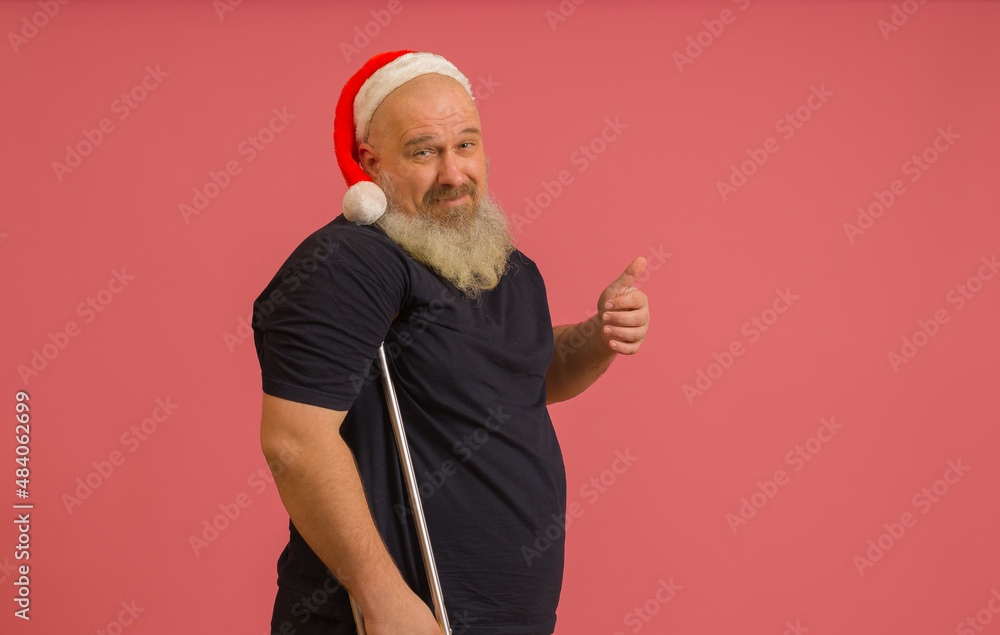 gray-haired man in black t-shirt and santa claus hat leaning on crutch shows thumb up on pink background